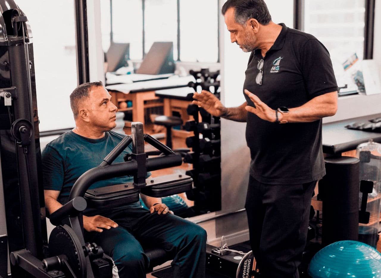 Profissional da MG conversando com uma pessoa sentada, em sala com equipamentos ao fundo.